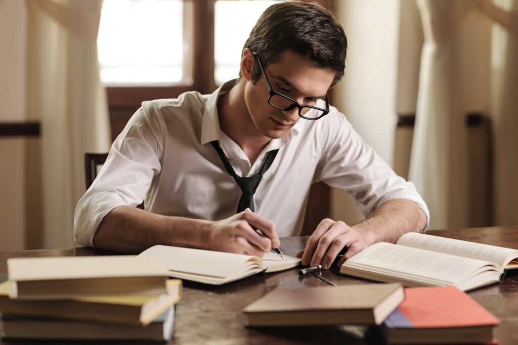 A man in his twenties, wearing glasses, sits at a desk, writing, surrounded by books. He is studying.