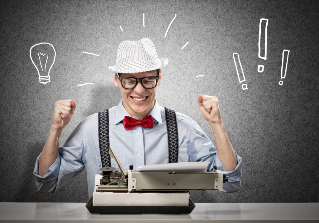 A man in his thirties, waering glasses and a hat, sits at a typewriter. He is smiling and triumphantly waving his fists in the air. He is having a lightbulb moment, as shown with an illustration of a lightbulb and three exclamation marks either side of his head.