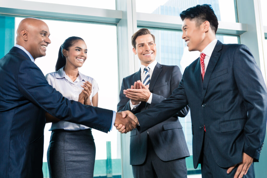 Two smartly dressed businessmen shaking hands in an office building whilst their female & male colleagues applaud them. All four are of mixed ethnicities.