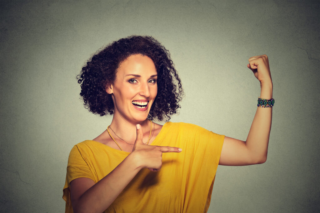 A picture of a young woman smiling and pointing at her curled bicep in an expression of inner power. 