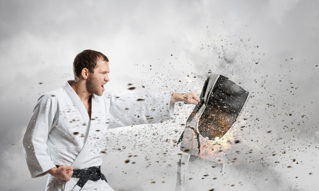 A man dressed in a white martial arts outfit and a black belt is energetically punching an inanimate object that is shattering around him from the impact.